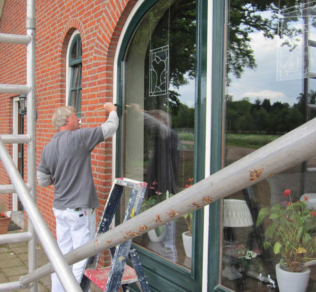 Schilder schildert een groen, rond kozijn aan een bakstenen woning, met een steiger ervoor